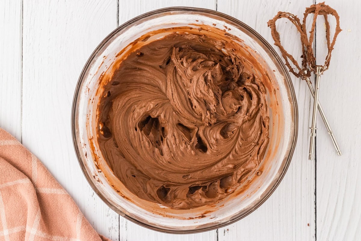 A glass bowl with fully mixed chocolate frosting, smooth and fluffy, with a hand mixer coated in frosting resting beside it.