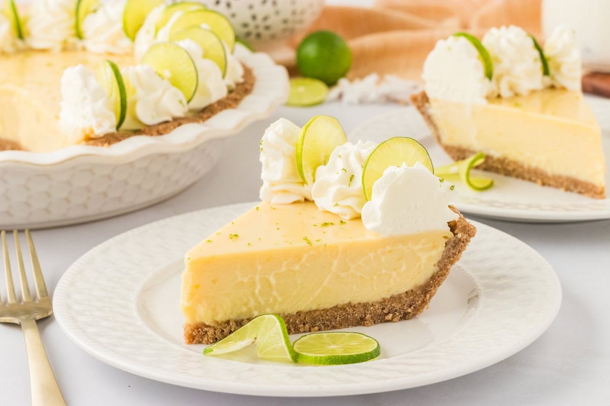 A plated slice of key lime pie with whipped cream and lime slices, ready to be served. Another slice and the whole pie are visible in the background.