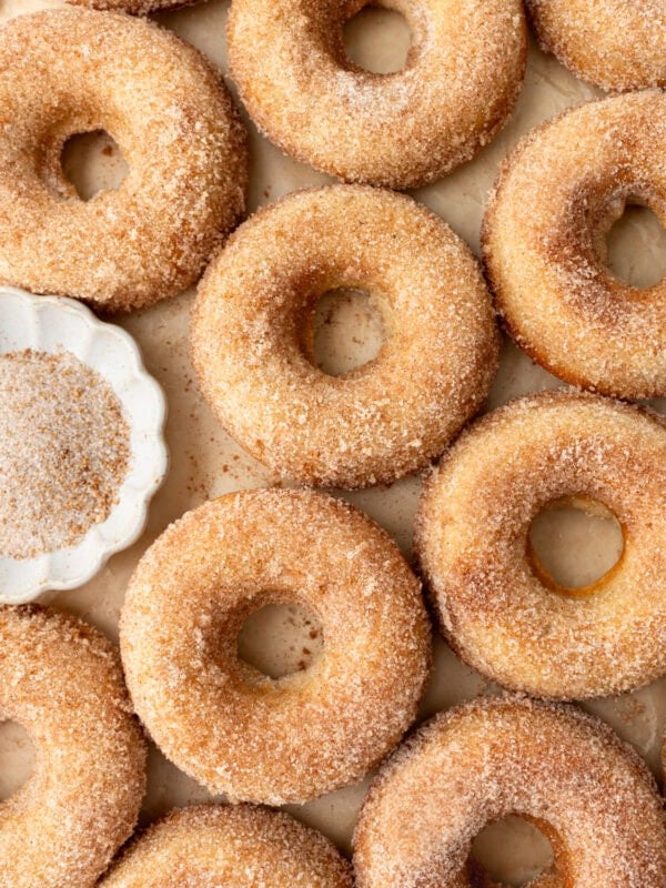 A close-up of freshly baked gluten-free cinnamon sugar donuts arranged on a parchment-lined surface, showing their golden brown texture with a light dusting of sugar. A small dish of cinnamon sugar sits nearby.