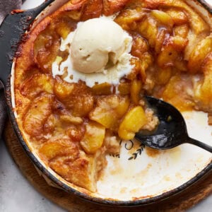 A close-up of a baked peach cobbler in a black skillet with a scoop of melting vanilla ice cream on top. The golden peaches and caramelized edges create a warm and inviting texture.