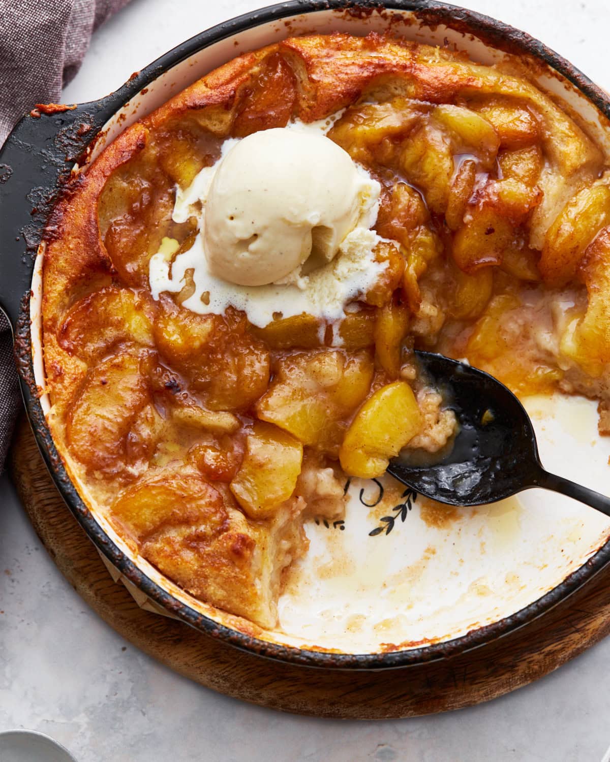 A close-up of a baked peach cobbler in a black skillet with a scoop of melting vanilla ice cream on top. The golden peaches and caramelized edges create a warm and inviting texture.
