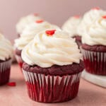 Close-up of a gluten-free red velvet cupcake with creamy swirled cream cheese frosting, topped with a small red heart decoration. Several cupcakes are in the background, slightly blurred.