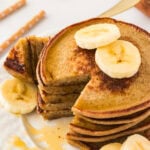 A close-up of a stack of banana oatmeal pancakes on a white plate, topped with fresh banana slices and drizzled with maple syrup, with a piece cut out to reveal the fluffy interior.
