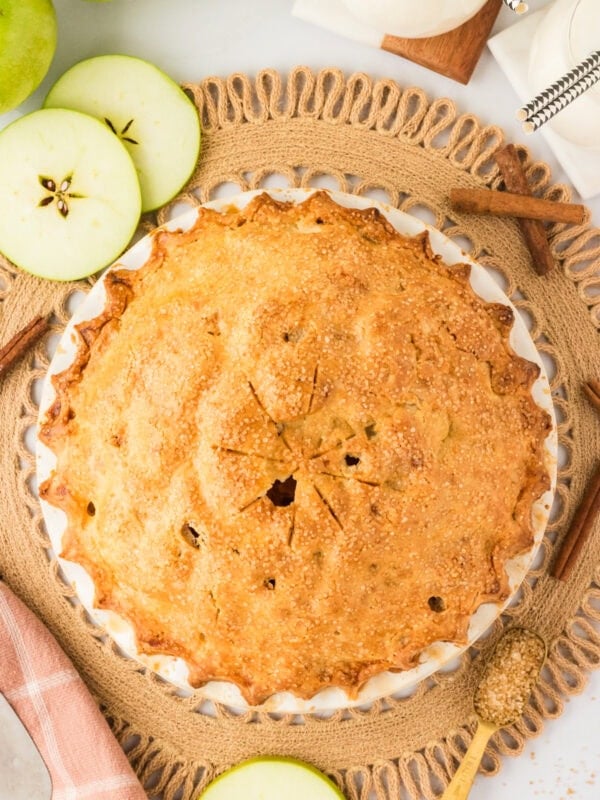 A golden-brown gluten-free apple pie with a flaky crust sits in a white pie dish, sprinkled with coarse sugar. The pie is surrounded by sliced green apples, cinnamon sticks, and a rustic woven placemat.