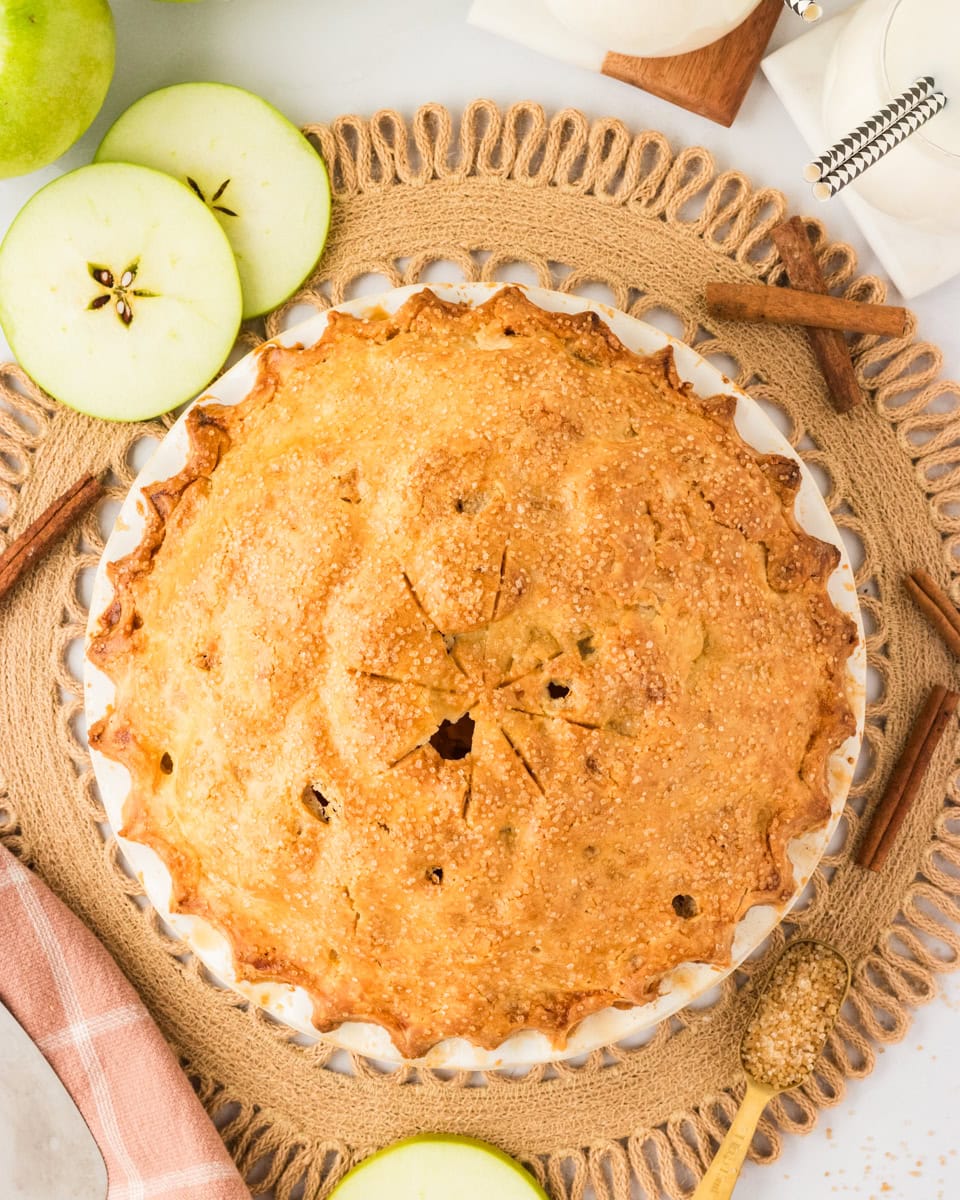A golden-brown gluten-free apple pie with a flaky crust sits in a white pie dish, sprinkled with coarse sugar. The pie is surrounded by sliced green apples, cinnamon sticks, and a rustic woven placemat.