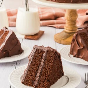 A slice of rich, two-layer chocolate cake with creamy chocolate frosting, served on a white plate. A full cake sits on a wooden cake stand in the background, with additional slices plated and glasses of milk nearby.