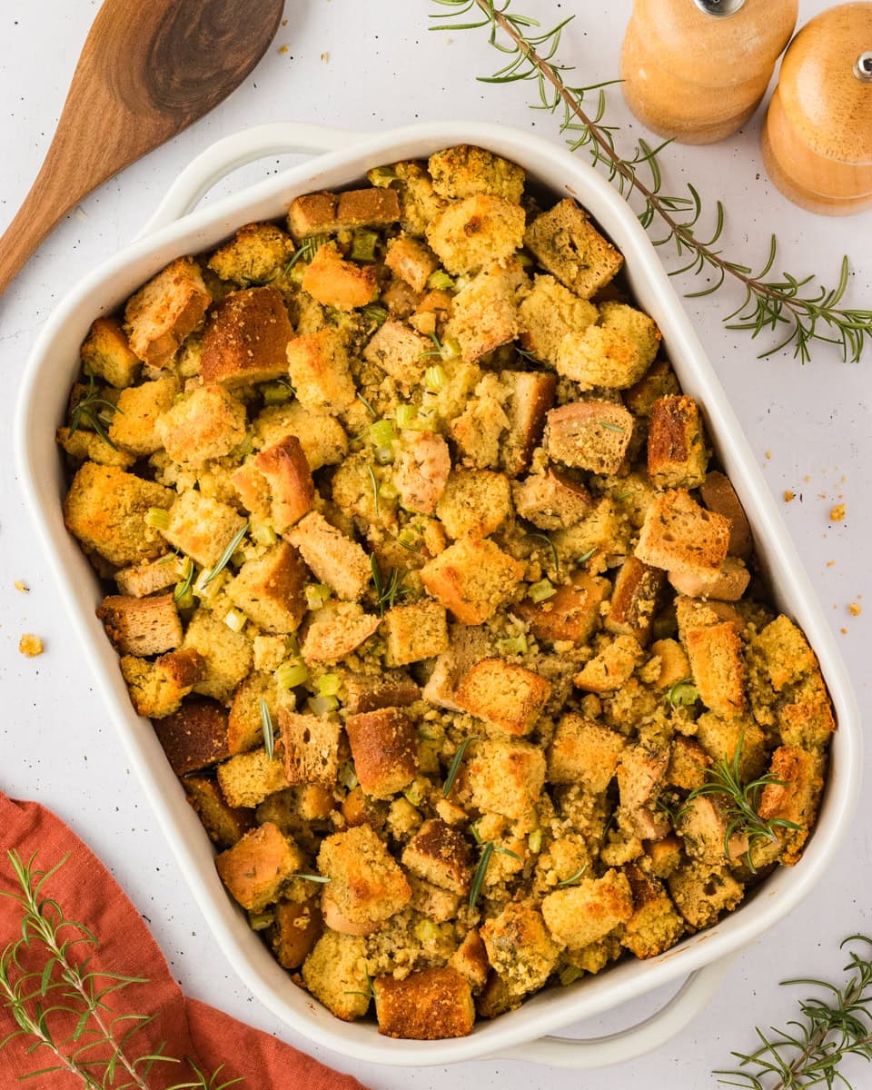 A baked dish of golden-brown gluten-free cornbread stuffing with herbs, served in a white casserole dish with a wooden spoon and fresh rosemary nearby.
