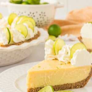 A slice of creamy gluten-free key lime pie on a white plate, topped with whipped cream and lime slices. A whole pie with additional slices is visible in the background, along with fresh limes in a white colander.