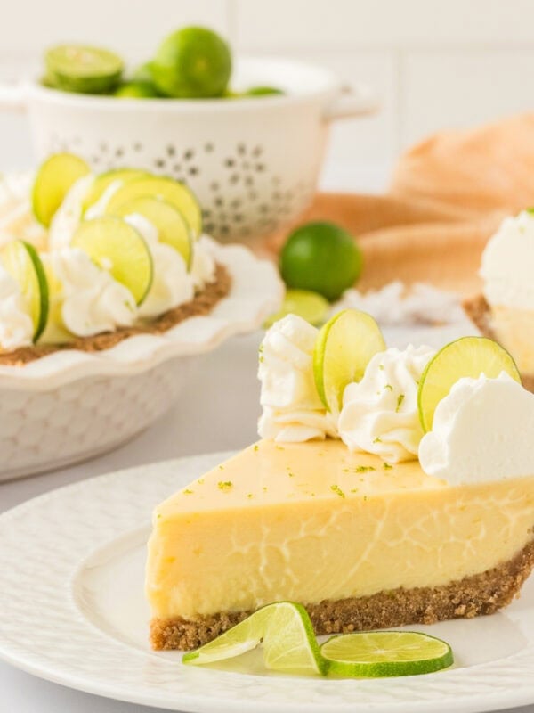A slice of creamy gluten-free key lime pie on a white plate, topped with whipped cream and lime slices. A whole pie with additional slices is visible in the background, along with fresh limes in a white colander.