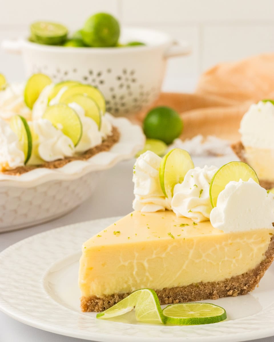 A slice of creamy gluten-free key lime pie on a white plate, topped with whipped cream and lime slices. A whole pie with additional slices is visible in the background, along with fresh limes in a white colander.