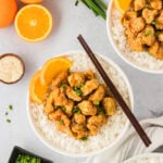 A white bowl filled with gluten-free orange chicken served over white rice, garnished with fresh orange slices, green onions, and sesame seeds. Whole oranges and scallions are displayed in the background for a fresh presentation.