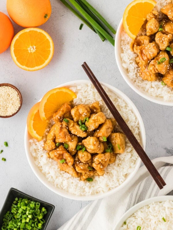 A white bowl filled with gluten-free orange chicken served over white rice, garnished with fresh orange slices, green onions, and sesame seeds. Whole oranges and scallions are displayed in the background for a fresh presentation.