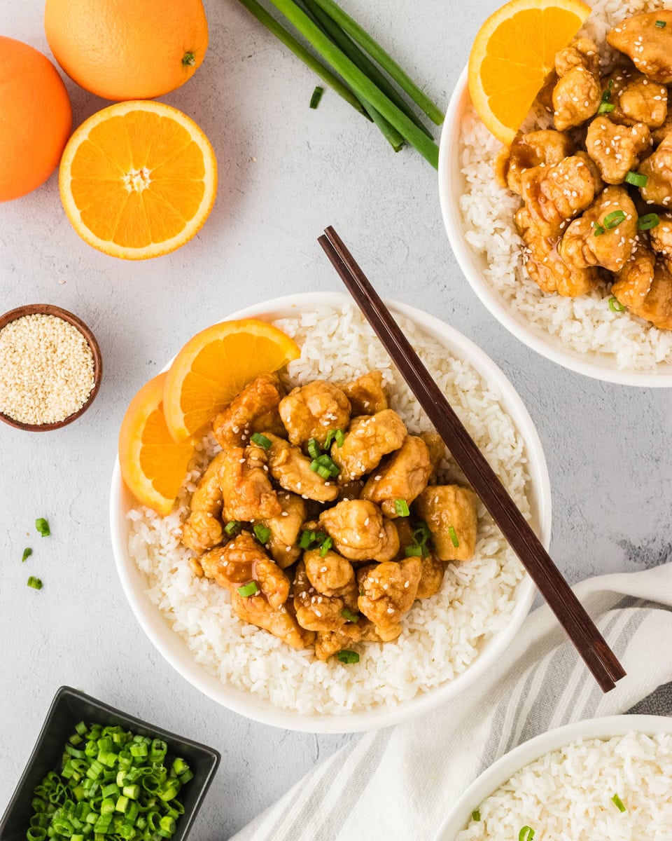 A white bowl filled with gluten-free orange chicken served over white rice, garnished with fresh orange slices, green onions, and sesame seeds. Whole oranges and scallions are displayed in the background for a fresh presentation.