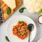 A white plate filled with golden brown cauliflower gnocchi covered in tomato sauce, garnished with fresh basil and grated cheese. A black spoon rests on the plate, surrounded by ingredients like Parmesan, fresh basil, and a head of cauliflower.