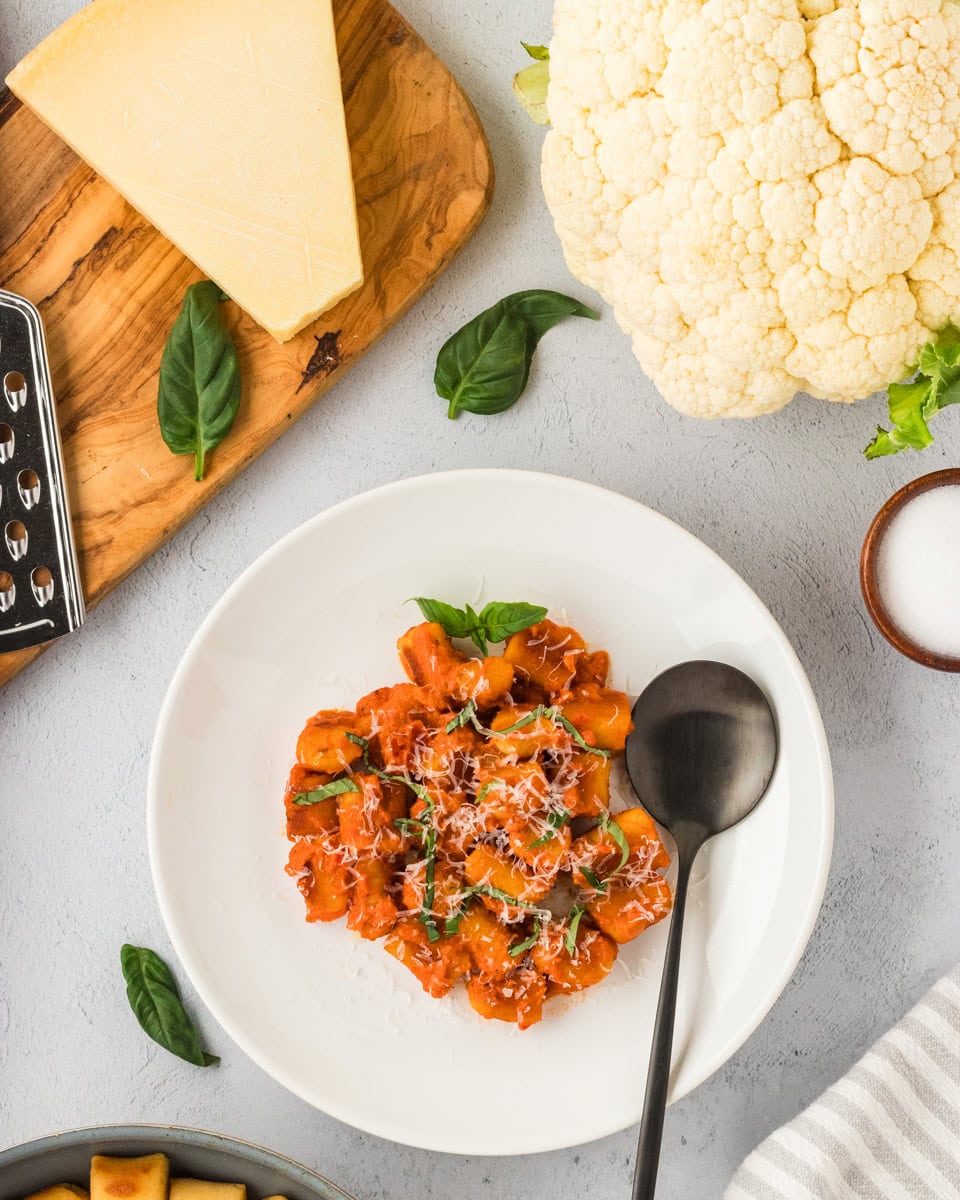 A white plate filled with golden brown cauliflower gnocchi covered in tomato sauce, garnished with fresh basil and grated cheese. A black spoon rests on the plate, surrounded by ingredients like Parmesan, fresh basil, and a head of cauliflower.