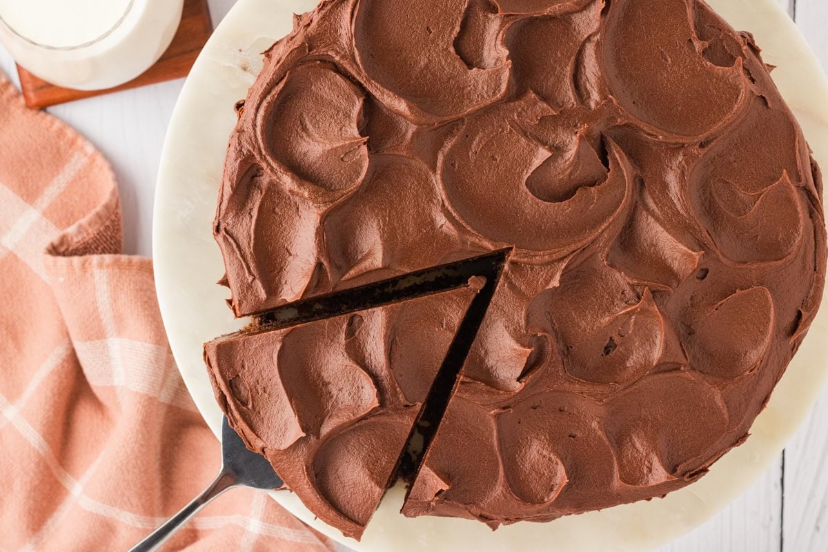 A slice being lifted from a fully frosted gluten-free chocolate cake, revealing its moist, dark layers beneath the thick chocolate frosting.