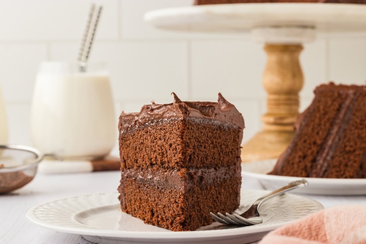 A close-up of a plated slice of gluten-free chocolate cake, showing two fluffy layers separated by creamy chocolate frosting, with glasses of milk and a wooden cake stand in the background.