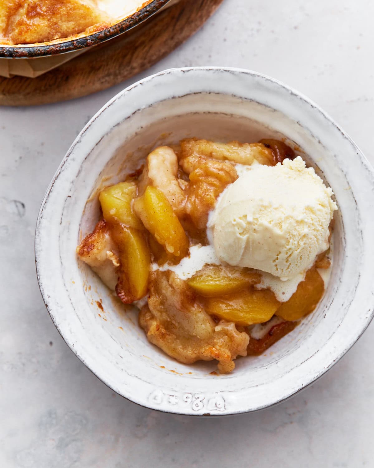 A serving of peach cobbler in a white bowl, topped with a scoop of vanilla ice cream. The cobbler has juicy peaches and a soft, golden crust.