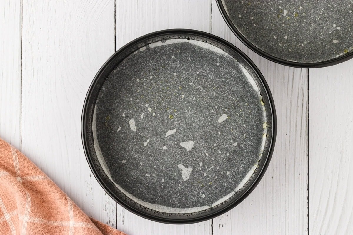Two round cake pans lined with parchment paper and lightly greased, sitting on a white wooden surface with a soft pink-checkered kitchen towel.