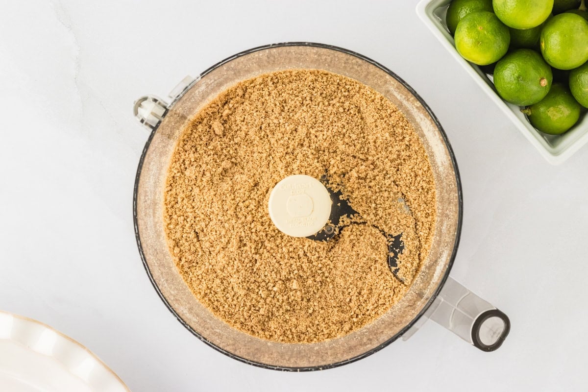 A close-up of a food processor filled with finely crushed Gluten-Free Cinnamon Chex, sitting next to a bowl of fresh limes.