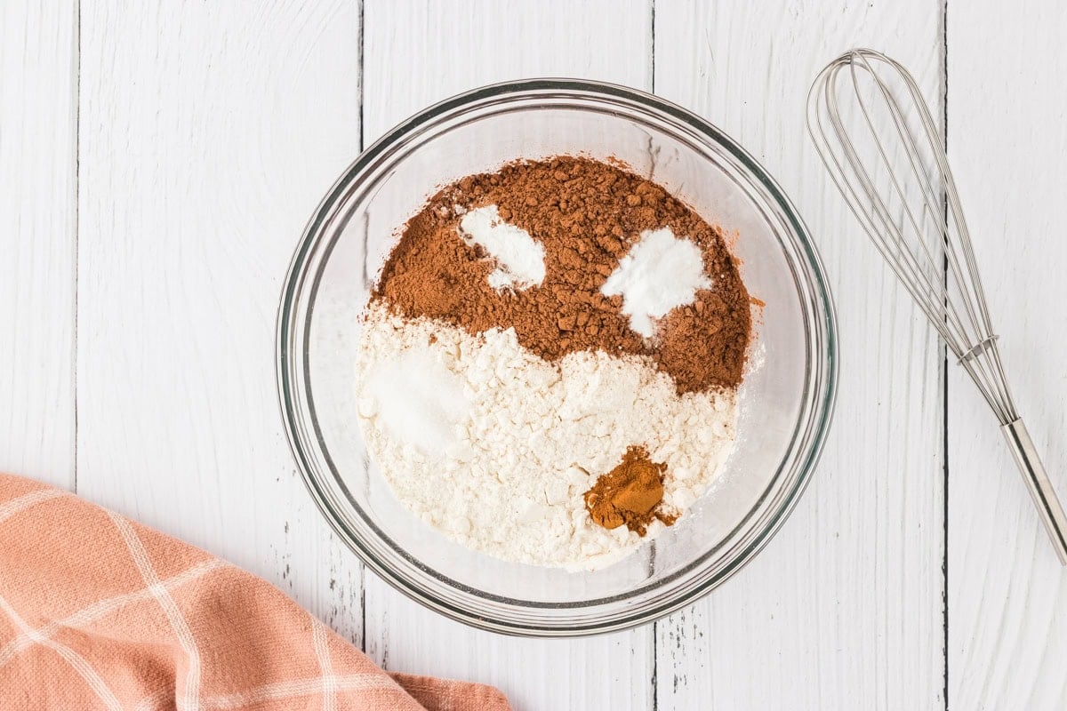 A glass mixing bowl containing dry ingredients for gluten-free chocolate cake, including cocoa powder, flour, baking soda, sugar, and cinnamon, with a whisk beside it.