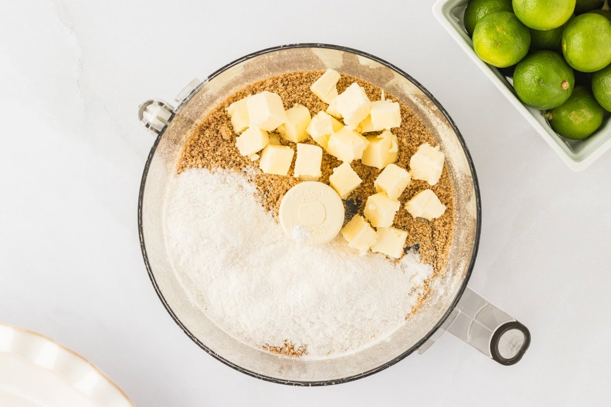 A food processor containing crushed Chex cereal, chopped unsalted butter, and Gluten-Free Bisquick, ready to be blended into a crust.