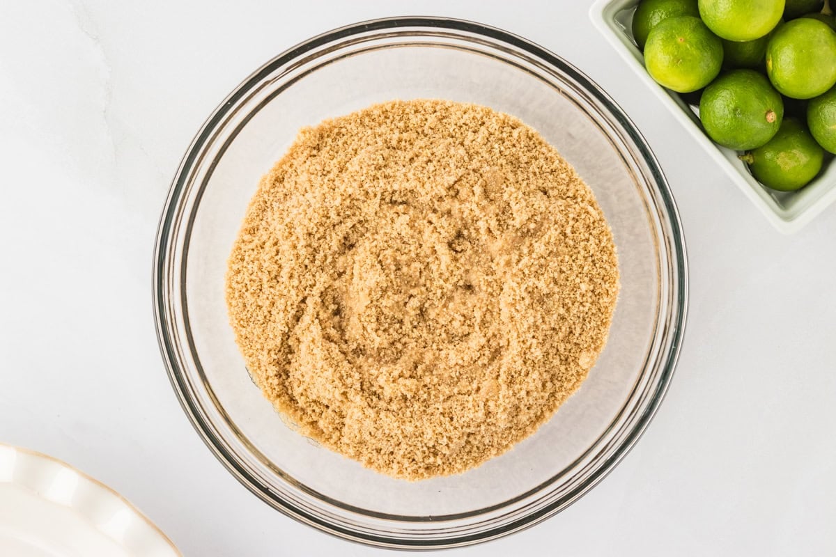 A medium glass bowl filled with the blended Gluten-Free Chex crust mixture, placed on a white countertop next to a bowl of fresh limes.
