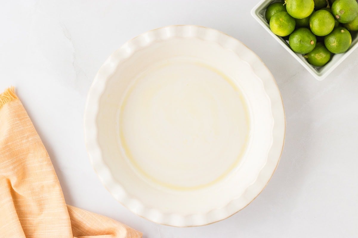 A 9-inch pie dish lightly coated with gluten-free coconut oil spray, set next to fresh limes and a peach-colored kitchen towel.