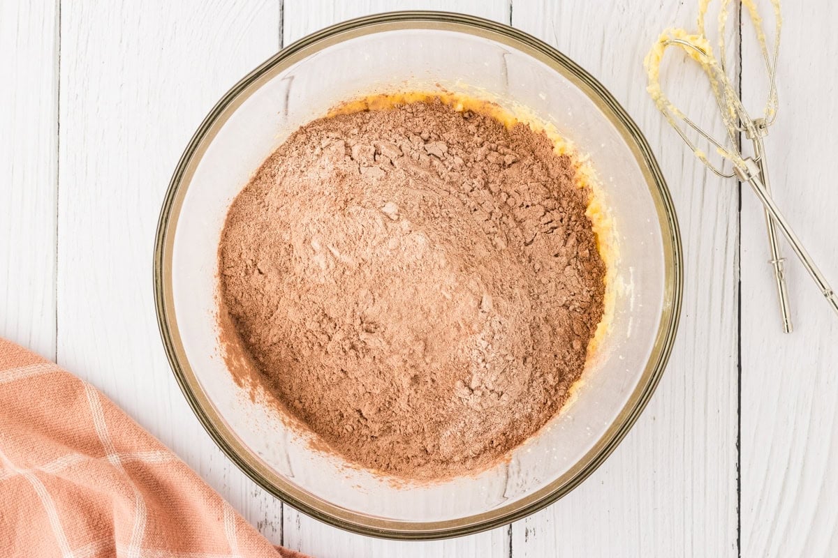 The dry cocoa-flour mixture added on top of the wet butter-egg mixture in a glass bowl, with a hand mixer nearby.