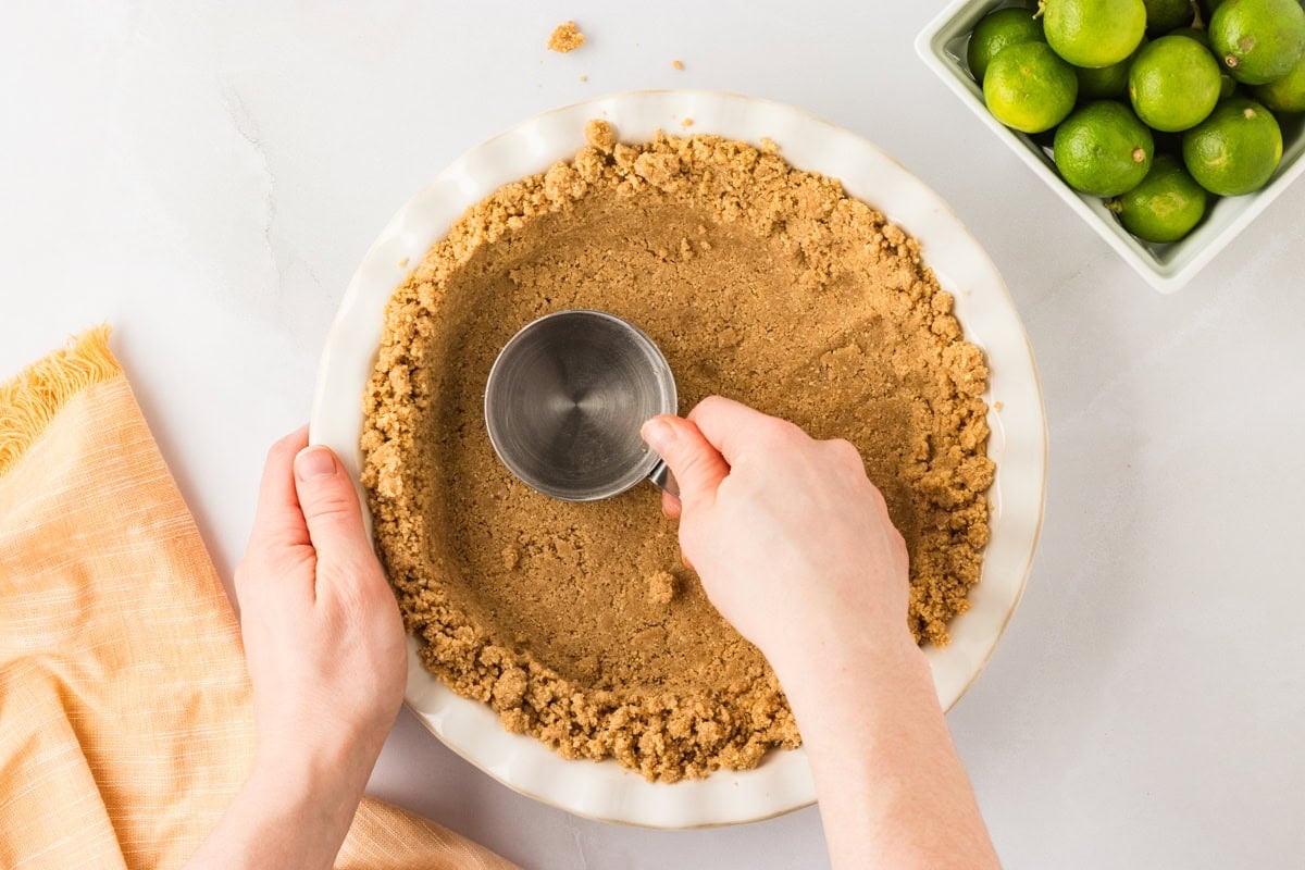 A close-up of hands pressing the Chex crust mixture into a pie dish using the bottom of a flat measuring cup to create an even surface.