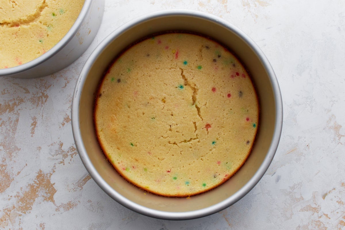 Two golden brown gluten-free funfetti cake layers in their pans, slightly domed with a few cracks on the surface, cooling before frosting.
