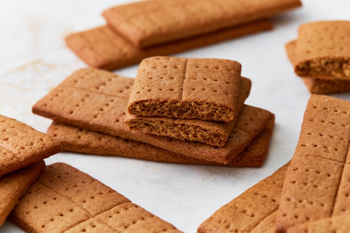 A close-up of a stack of gluten-free graham crackers, with one broken in half to reveal its crunchy texture inside. The crackers have a rich golden-brown hue with visible fork pricks.