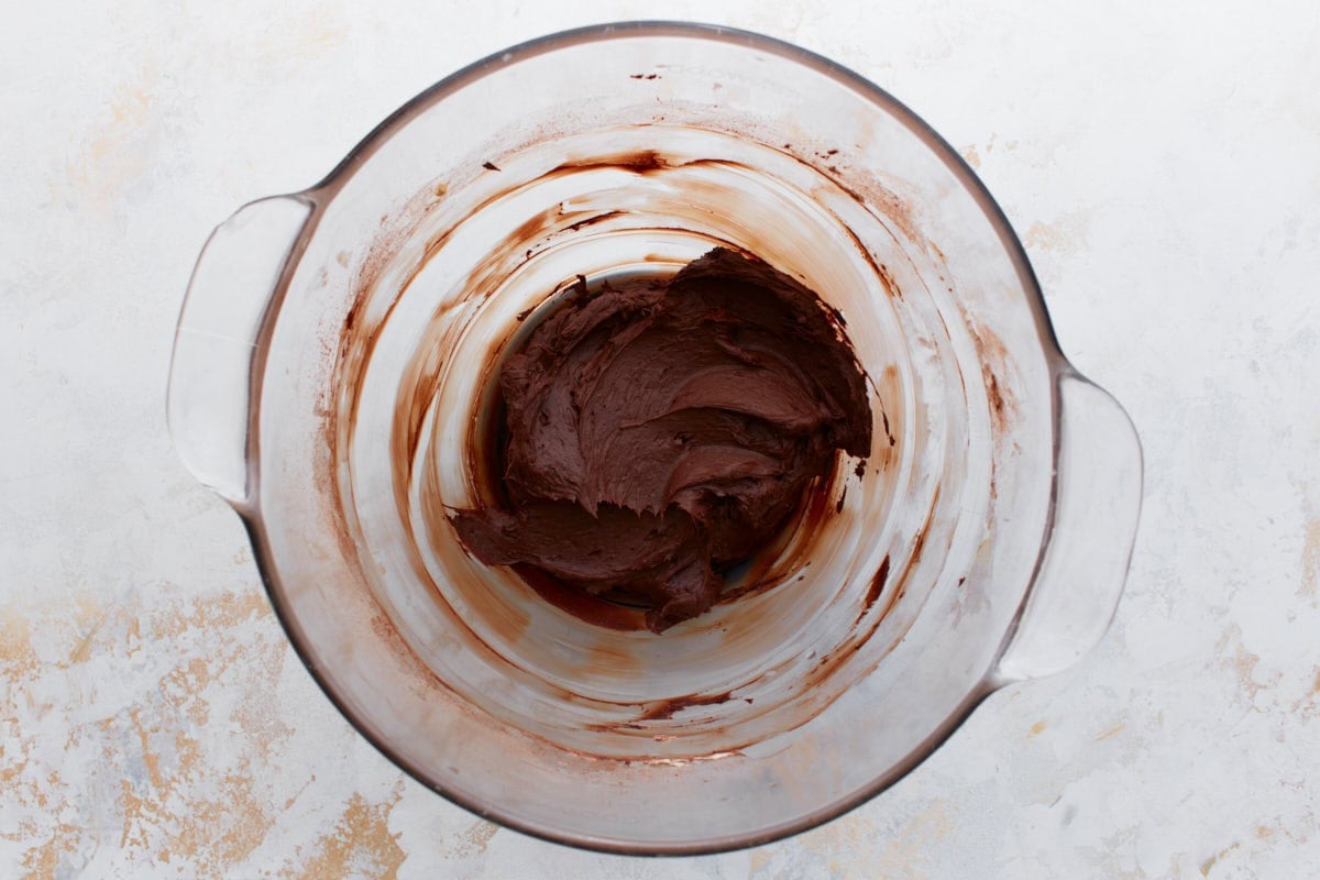 A glass bowl with thick chocolate buttercream, partially mixed with visible cocoa powder streaks.