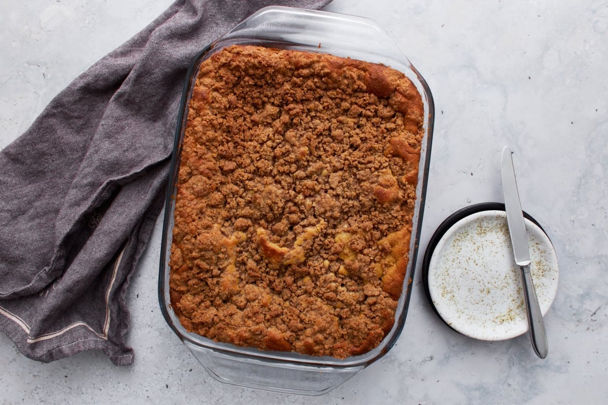 A full view of the baked coffee cake in a glass dish, resting on a countertop with a gray linen napkin and an empty plate with a knife beside it.