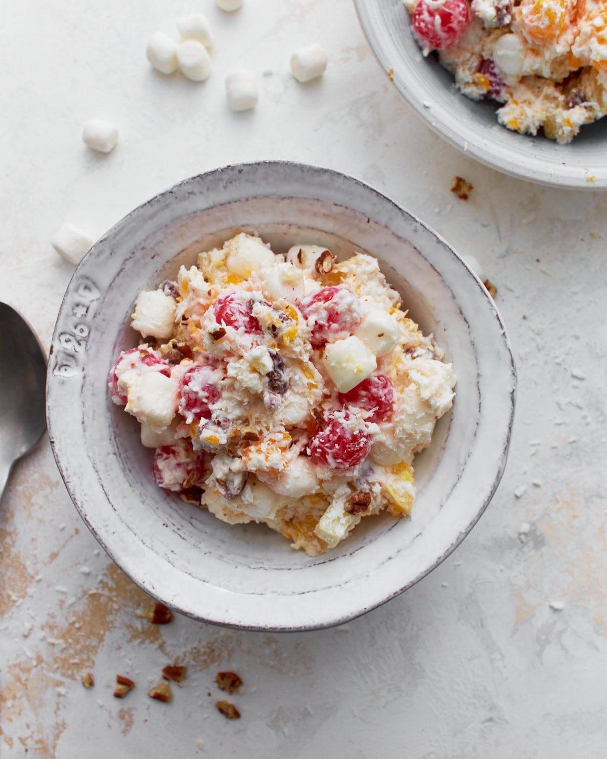 A bowl of ambrosia salad filled with creamy whipped topping, pineapple chunks, mandarin oranges, maraschino cherries, mini marshmallows, shredded coconut, and chopped nuts.