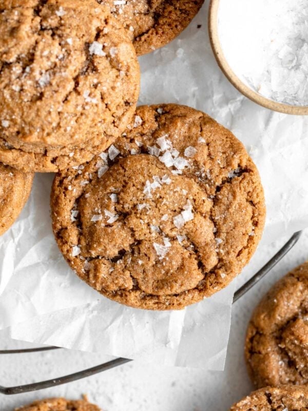A close-up of golden-brown flourless peanut butter cookies topped with flaky sea salt, resting on crinkled parchment paper.