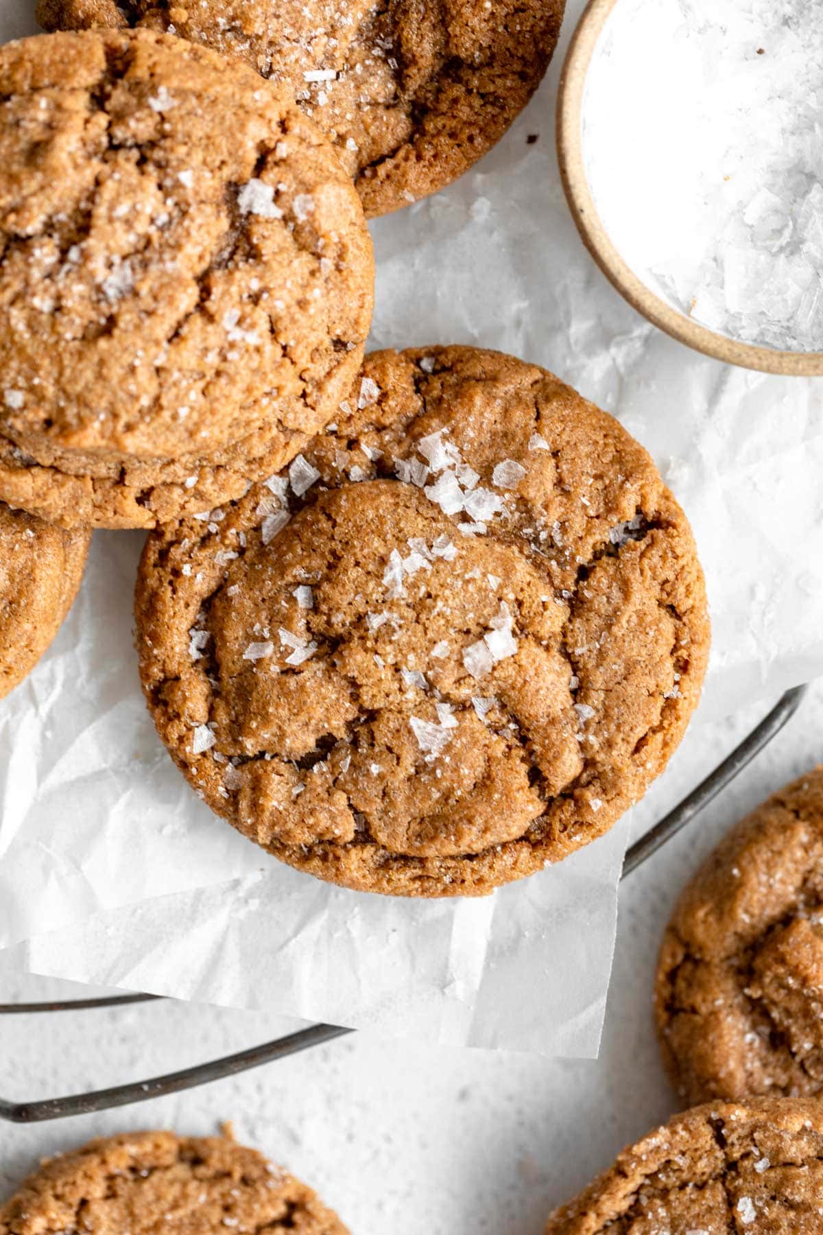 A close-up of golden-brown flourless peanut butter cookies topped with flaky sea salt, resting on crinkled parchment paper.