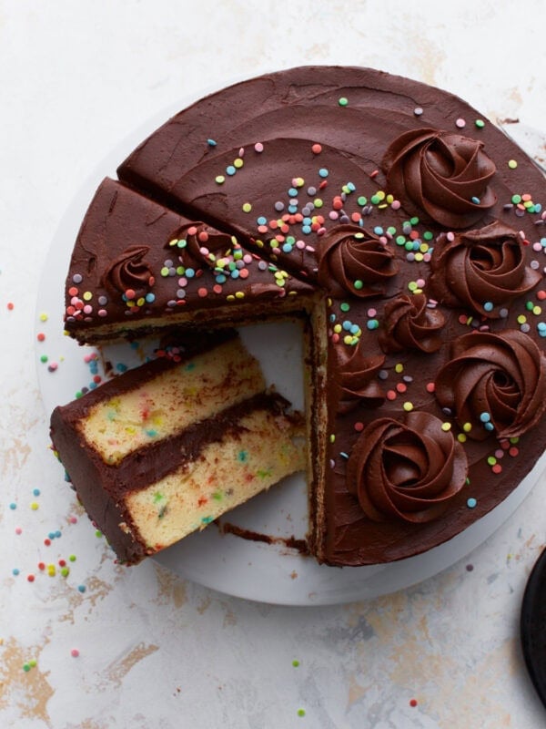 A gluten-free funfetti birthday cake covered in rich chocolate buttercream, decorated with piped swirls and colorful sprinkles, with a slice removed to reveal the soft yellow cake with sprinkles inside.