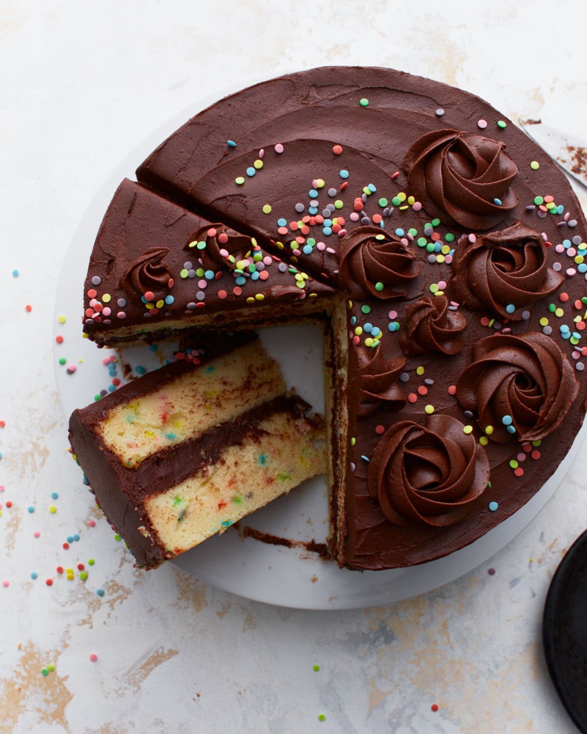 A gluten-free funfetti birthday cake covered in rich chocolate buttercream, decorated with piped swirls and colorful sprinkles, with a slice removed to reveal the soft yellow cake with sprinkles inside.