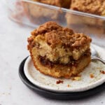 A close-up of a single slice of gluten-free cinnamon coffee cake on a white plate with a black rim, showing a rich cinnamon swirl and crumbly streusel topping. A glass baking dish with more cake is visible in the background.