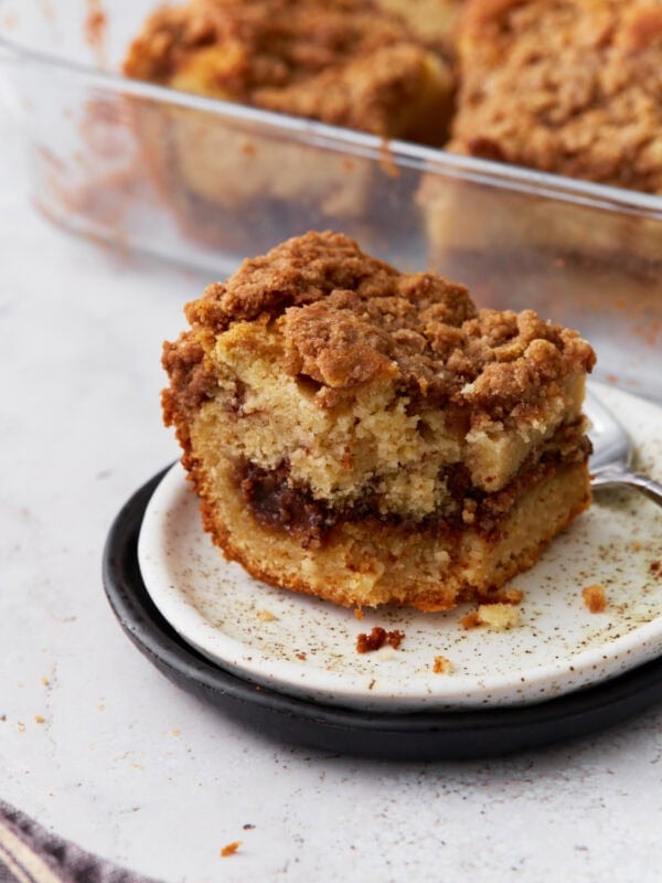 A close-up of a single slice of gluten-free cinnamon coffee cake on a white plate with a black rim, showing a rich cinnamon swirl and crumbly streusel topping. A glass baking dish with more cake is visible in the background.