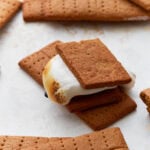 A close-up of a freshly made gluten-free s’more with a toasted marshmallow and chocolate between two homemade graham crackers. Several whole and broken graham crackers surround it on a white surface.