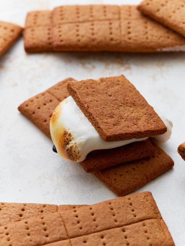 A close-up of a freshly made gluten-free s’more with a toasted marshmallow and chocolate between two homemade graham crackers. Several whole and broken graham crackers surround it on a white surface.