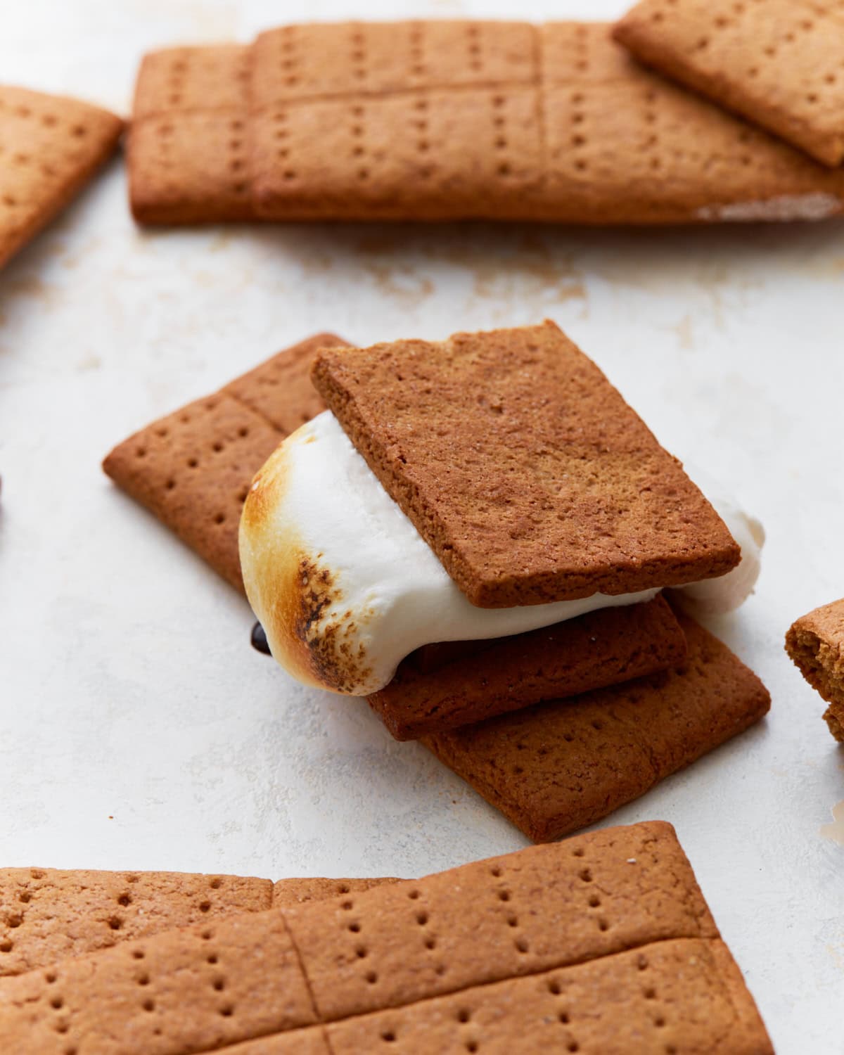 A close-up of a freshly made gluten-free s’more with a toasted marshmallow and chocolate between two homemade graham crackers. Several whole and broken graham crackers surround it on a white surface.