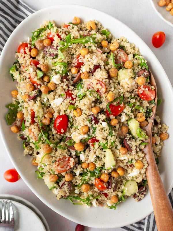 A white oval dish filled with Greek quinoa salad, featuring fluffy quinoa, cherry tomatoes, cucumbers, Kalamata olives, arugula, feta cheese, and crispy roasted chickpeas. A wooden spoon rests in the dish.