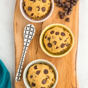 Three small bowls filled with edible chickpea cookie dough topped with chocolate chips, arranged on a wooden serving board. A decorative spoon and scattered chocolate chips are placed nearby.