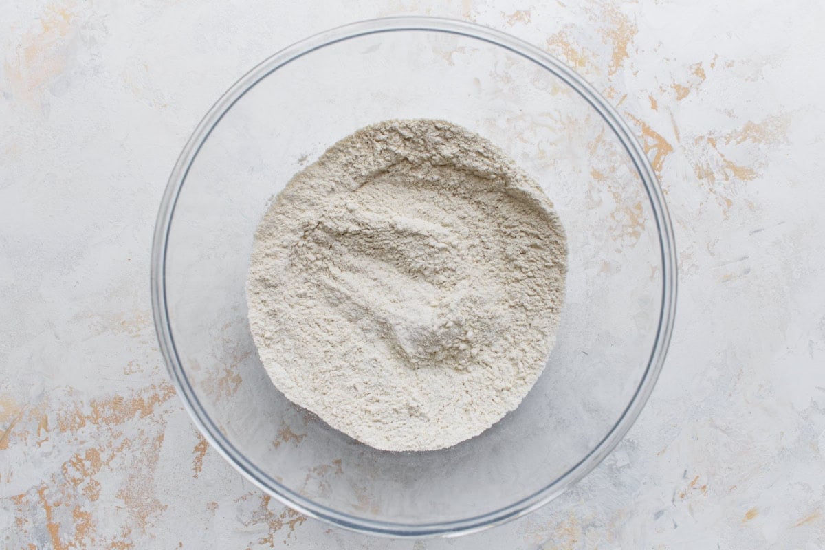 A glass mixing bowl filled with dry buckwheat flour on a textured light-colored surface, ready for pancake preparation.