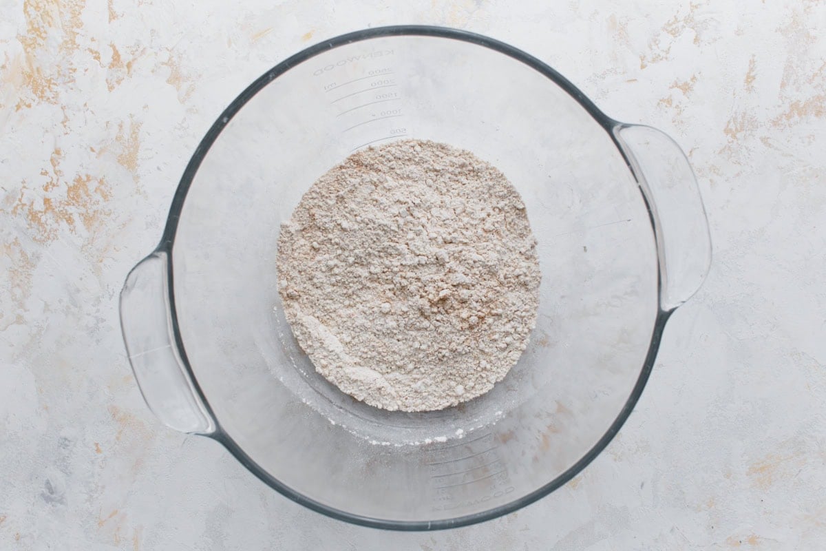 A glass mixing bowl filled with dry ingredients for gluten-free graham crackers. The mixture is light brown and appears well combined.