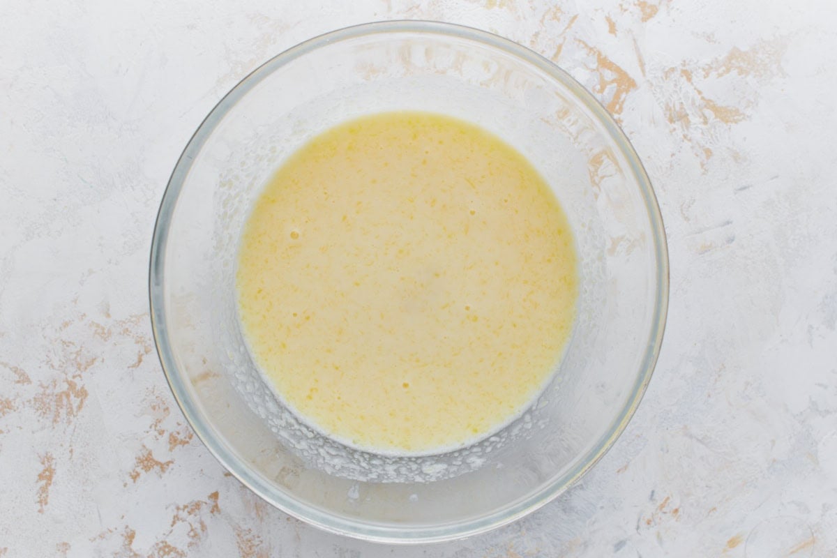 A glass bowl containing a whisked wet ingredient mixture with a yellow hue, likely including eggs, milk, and melted butter, for making buckwheat pancakes.