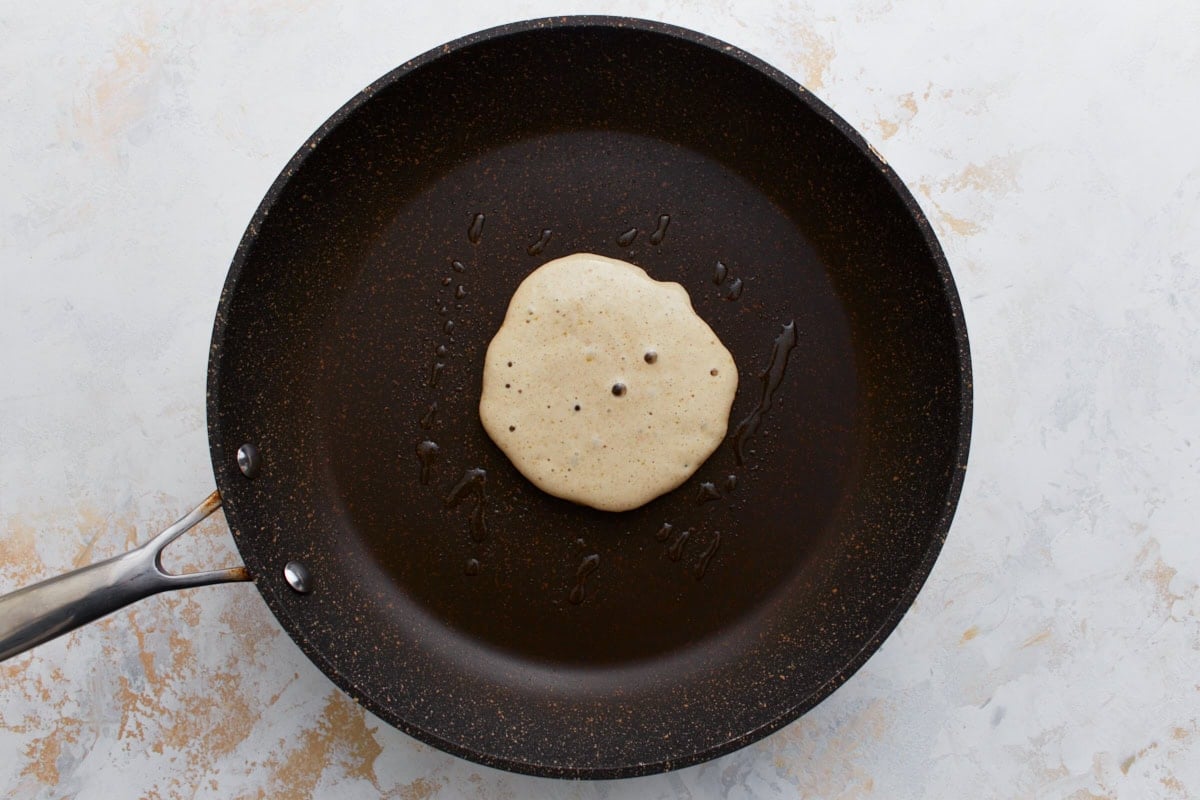 A single buckwheat pancake cooking in a lightly greased non-stick pan, with small bubbles forming on the surface, showing the early stage of cooking.